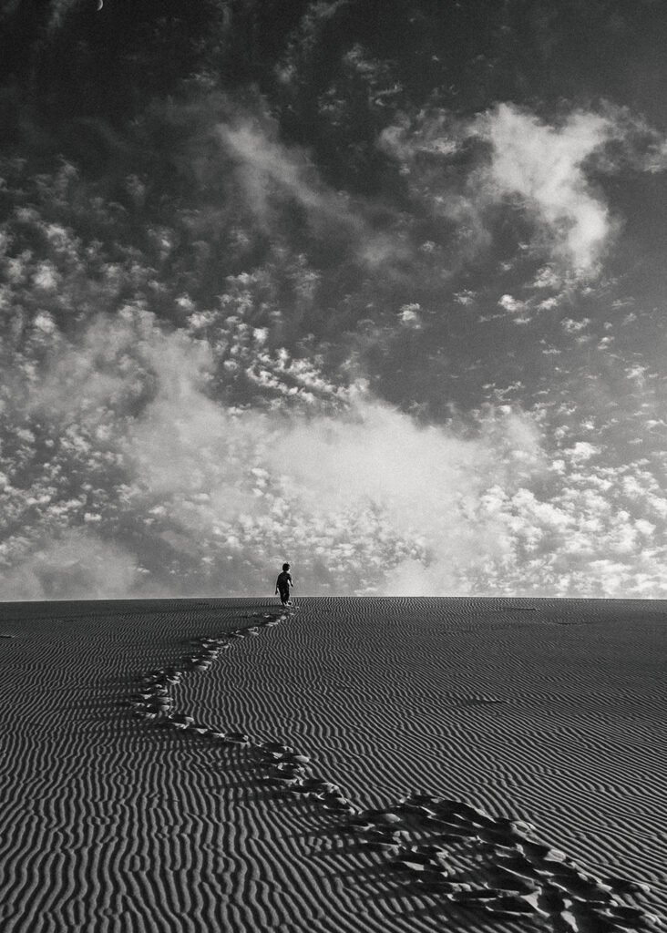 Black and white Environmental portrait of child climbing sand dune toward clouds