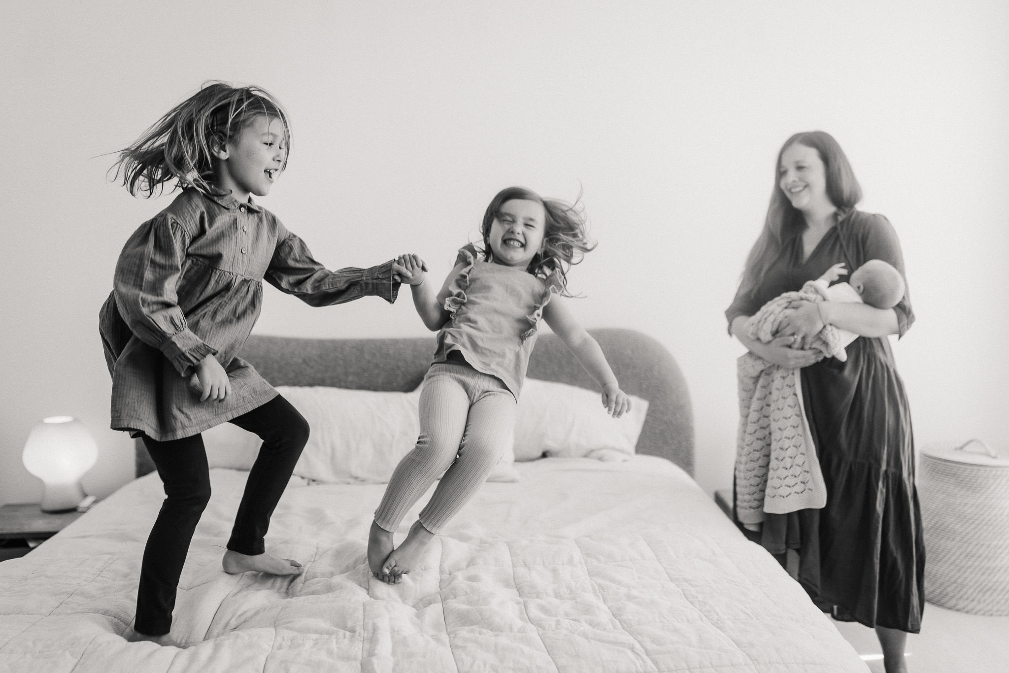 black and white photo of siblings jumping on bed at in home newborn session by Ashley Kaplan Photography